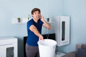 In an Atlanta living room, a man balances a white bucket under water streaming from the ceiling while calmly talking on the phone.
