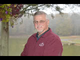 A person in a maroon jacket stands outdoors near a lake and trees, with a hanging plant visible to the left.