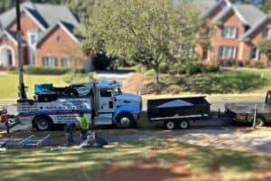 A dump truck and additional equipment, essential for the sewer repair in Vinings, are parked on a suburban street. Workers and materials are visible on the ground, with brick houses and trees forming a picturesque background.