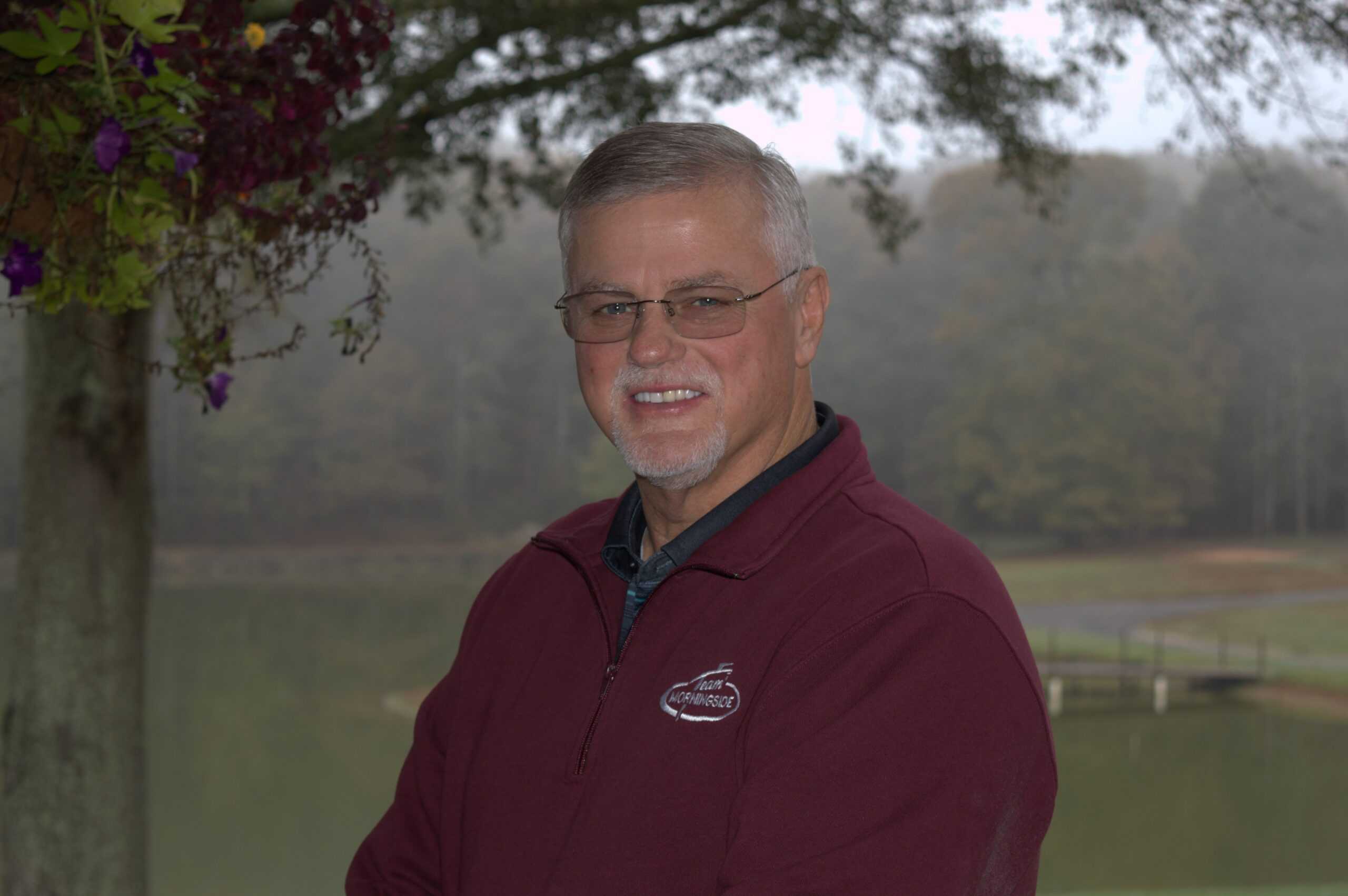 Man in a maroon sweater stands outdoors by a lake, smiling at the camera.