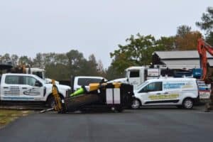 A horizontal directional drilling machine has collided with plumbing company vans near Midtown Atlanta. Several vehicles and an excavator are parked nearby on a paved area, with trees providing a scenic backdrop.