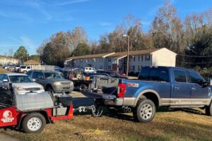 A blue pickup truck with a red and white trailer, possibly used for sewer jetting, is parked on a grassy area next to a sidewalk in Walnut Grove. Several other cars and a row of residential buildings are visible in the background.