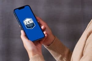 A person holds a smartphone displaying a blue screen with an "emergency plumber Brookhaven 24/7" logo.