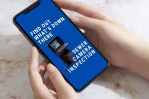 A person holds a smartphone displaying an ad for midtown Atlanta sewer camera inspections, featuring text and an image of a sewer camera on a blue background.